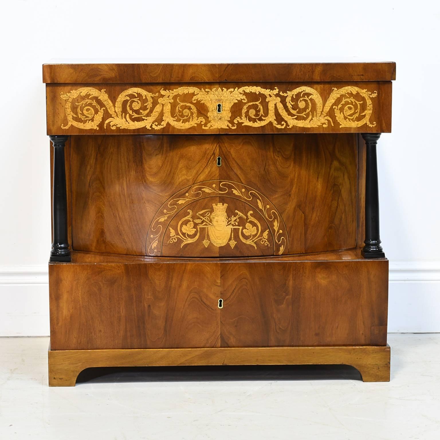 A very fine North German Biedermeier parcel-ebonized mahogany commode with top above a single drawer floridly inlaid with scrolling acanthus marquetry, the second convex-drawer inlaid with a scrolling foliate lunette and flanked by ebonized columns,