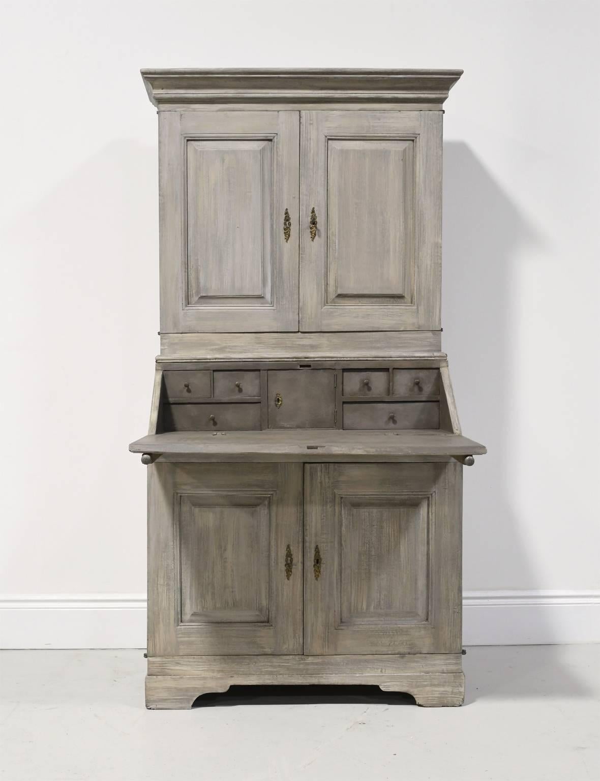 A very beautiful Gustavian secretary with fall-front opening to a desk offering six interior drawers surrounding a small cabinet door for valuables. A bookcase or cupboard rests over the desk with a cabinet below for added storage. The grey-painted