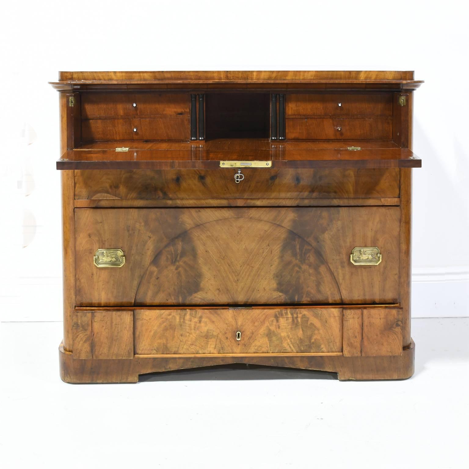 A Biedermeier chest in figured and bookmatched West Indies mahogany with three storage drawers of various depths below a drawer-front that opens to a secretary desk with four interior drawers around an open cubby, North German, circa