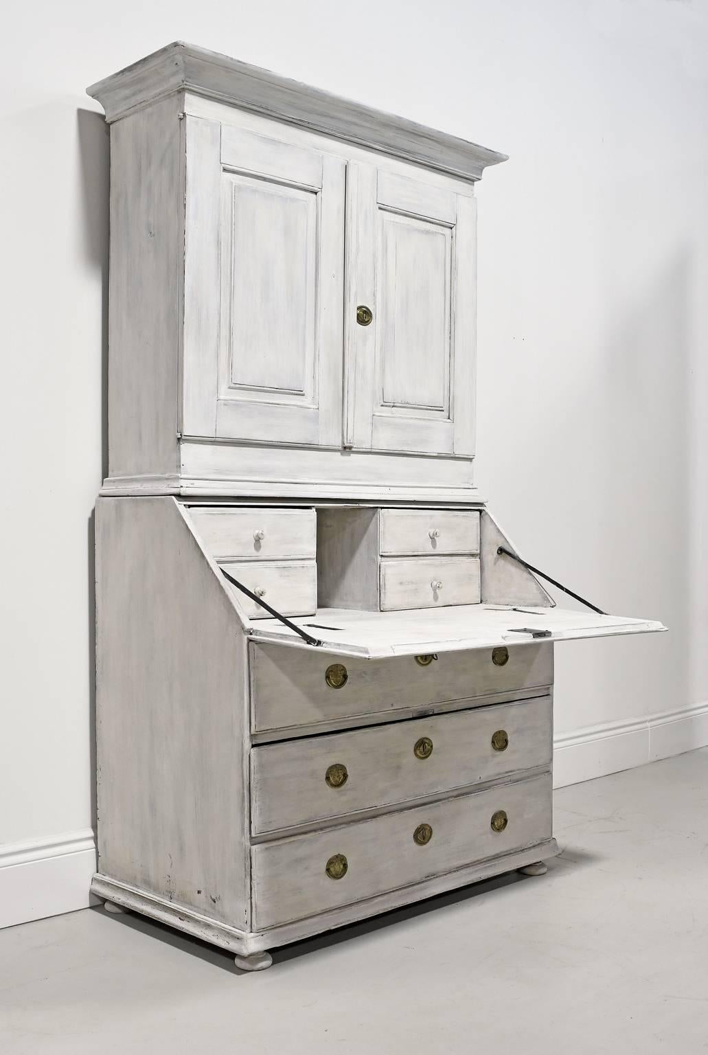 A rustic Swedish Gustavian 18th Century secretary in a grey white milk paint applied over multiple layers of previous finishes features a bookcase/cupboard seated on a fall-front, slant top secretary that opens to a desk with a cubby and four small