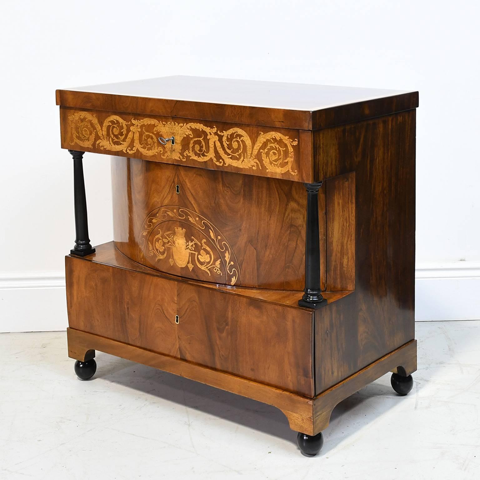 A very fine Biedermeier parcel-ebonized mahogany commode with top above a single drawer floridly inlaid with scrolling acanthus marquetry, the second convex-drawer inlaid with a scrolling foliate lunette and flanked by ebonized columns, and the