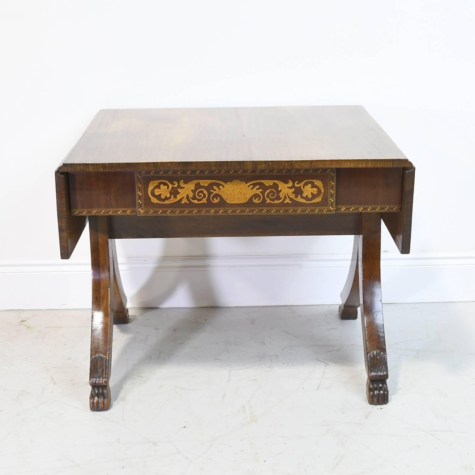 A handsome Italian Empire desk or sofa table in mahogany with fold-down leaves, and resting over trestle base with splayed legs ending in carved lions' paw feet. Offers one drawer decorated with marquetry inlays in an arabesque design, with the
