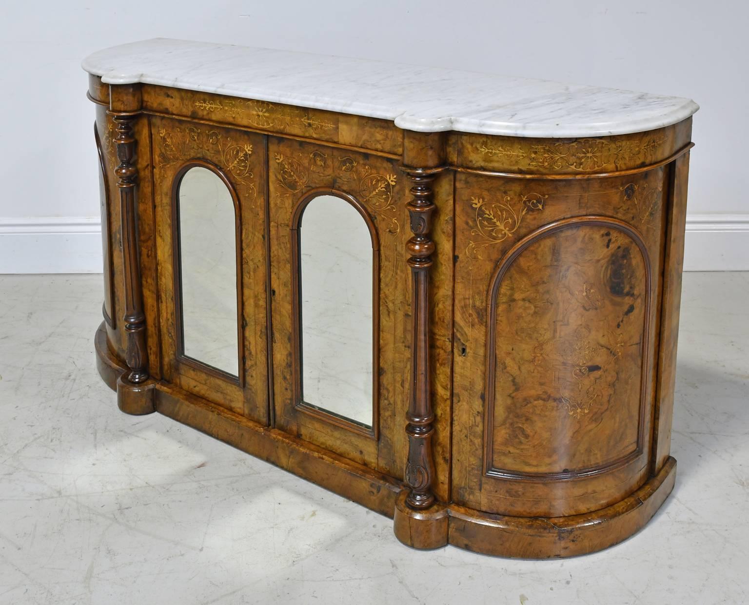 An early English Victorian console or sideboard in "D" form in burled walnut with marquetry inlays in satinwood, and white Carrara marble top. Features two centre cabinet doors with arched mirrors in panels, which are flanked by turned