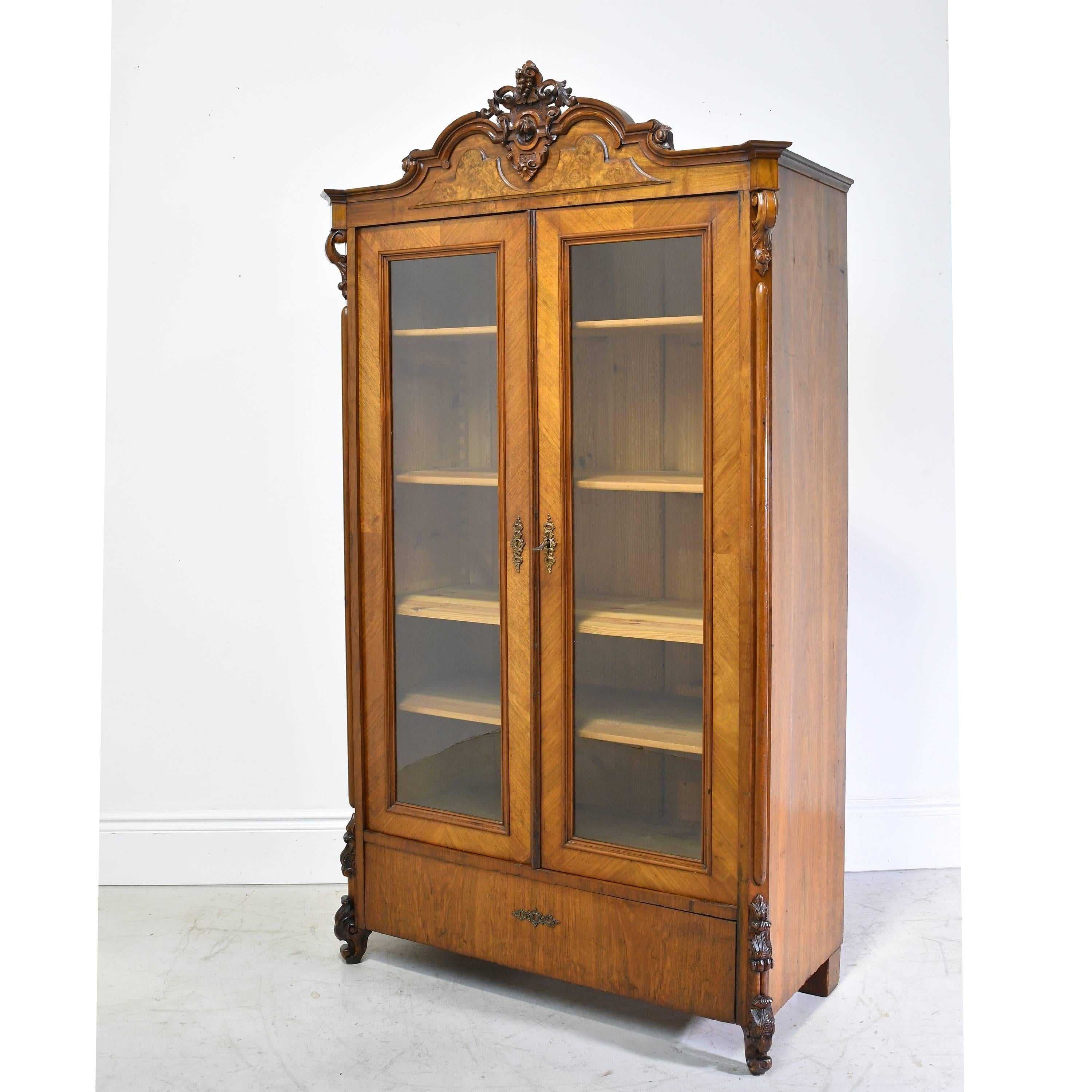A very lovely Louis Philippe bookcase or display case in walnut with carved bonnet over arched crest, two doors with glass panels, and resting on carved scroll feet. Has four adjustable shelves, original hardware with working lock on doors. Exterior