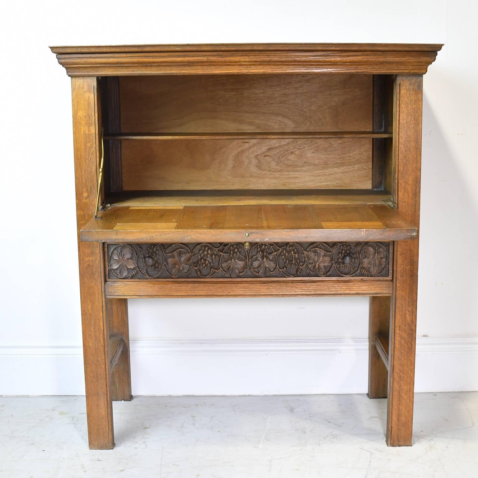 A handsome and rather singular Arts & Crafts bar cabinet in oak with fall-front and resting on square legs. Embellishments include carved linen-fold on the sides and front door panels that flank a centre panel of carved grapes and foliage that is