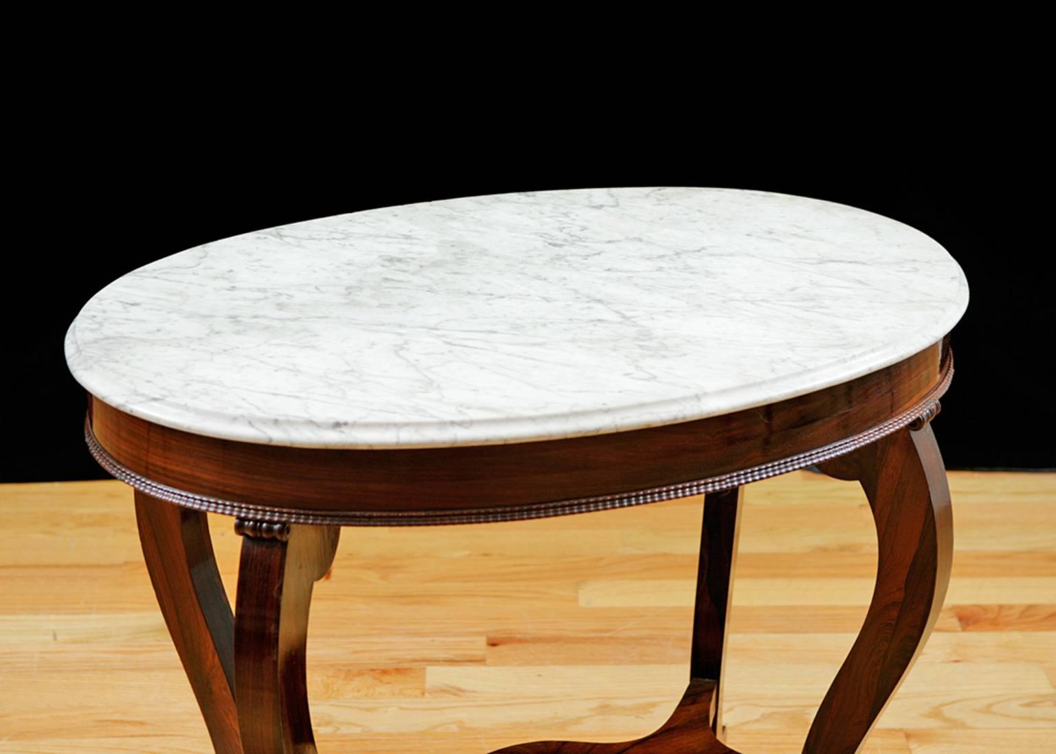 A graceful table with quiet refinement made of the finest rosewood, with the original white Carrara-marble top. Oval top with carved trim on apron is on cabriolet legs with scroll feet joined by a bottom stretcher, and rests on casters for mobility.