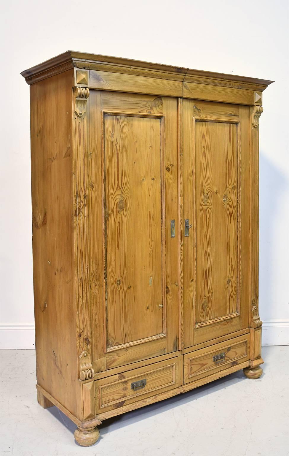 A handsome light, caramel-colored pine armoire with two doors with molding around the recessed panels that open on inside hinges to three fixed, interior shelves, with two exterior drawers on the base that rests on bun feet. Carved wooden appliqués