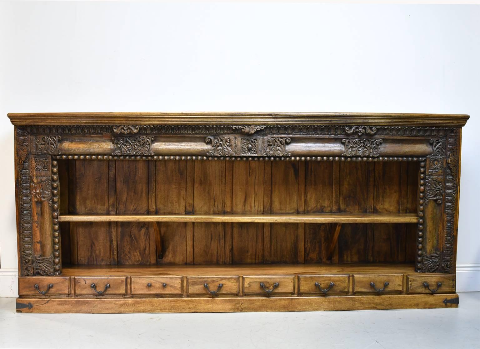 A long bookcase in teak made from antique carved architectural elements of Indian Himalayan influence with two shelves above a set of eight drawers that run horizontally along the bottom. Carved embellishments include acanthus leaves, lotus and