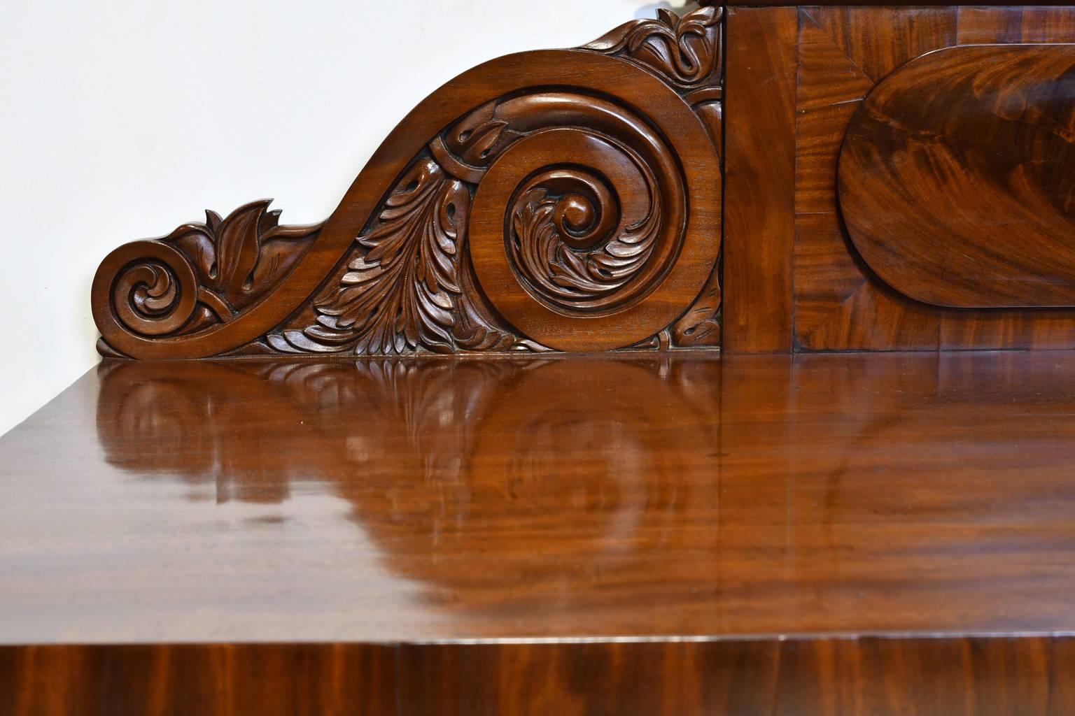 A fine and elegant English Regency pedestal sideboard in mahogany with carved backsplash. Offers three convex silverware drawers spanning the length just below the top, with shelving on one pedestal and two celarette drawers on the other for storage