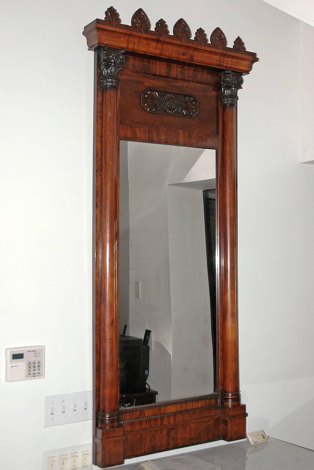 A very beautiful tall stately and elegant Empire mirror in fine Cuban mahogany with well-articulated carvings of anthemions as antefix decorating the crown molding. Turned columns with finely carved acanthus capitals flank the mirror & frieze.