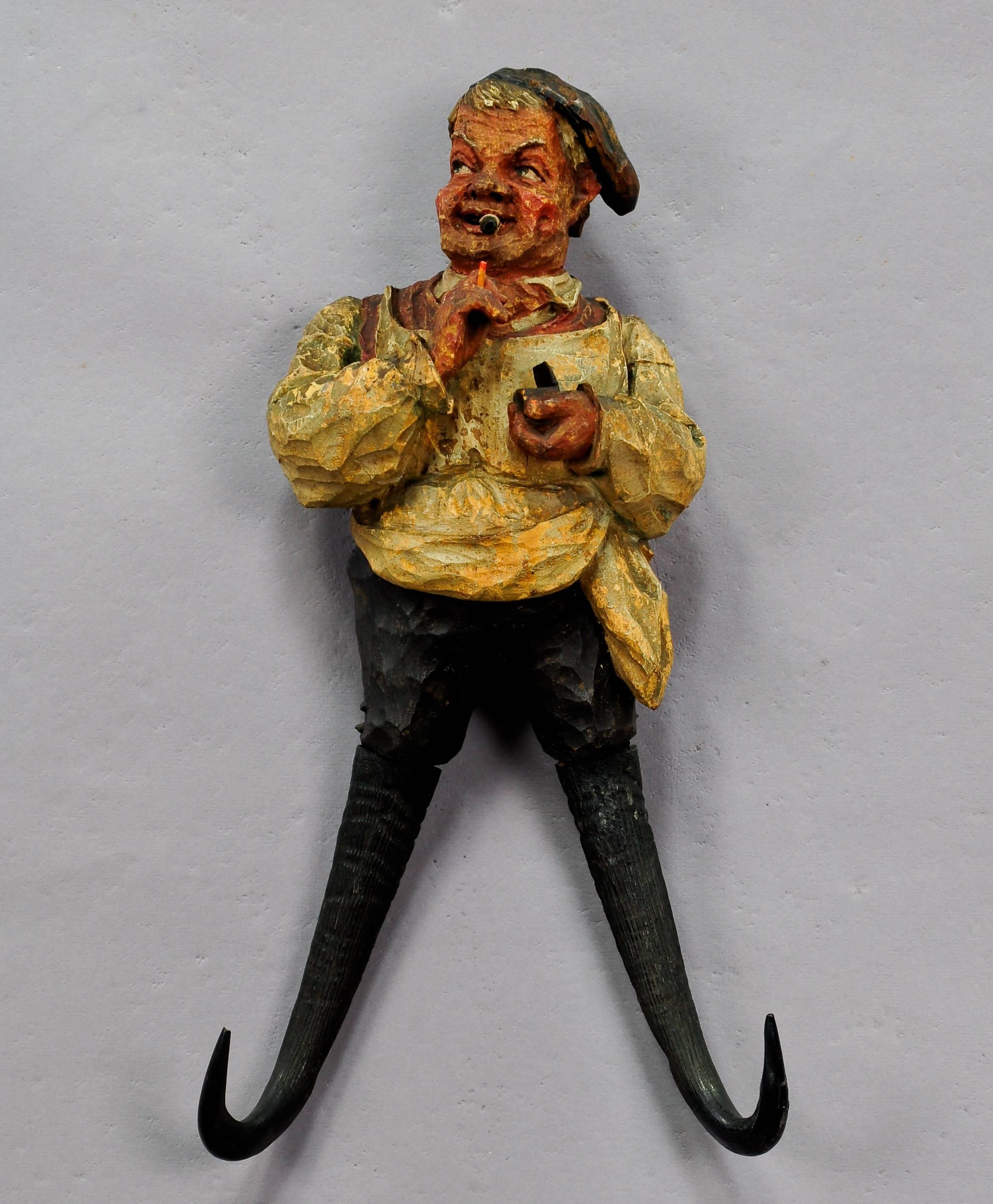 A whimsical black forest whip holder / coat hook carved as a farmer which is lighting a cigarette, circa 1920. With chamois horns, original paint with abrasions.