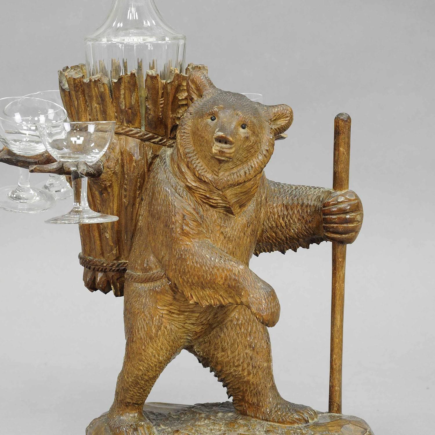 A carved linden wood decanter / liqueur stand modelled as hiking bear with walking stick and basket on his back. With decanter and six shot glasses. Executed, circa 1900, Swiss Brienz. (Height without bottle).