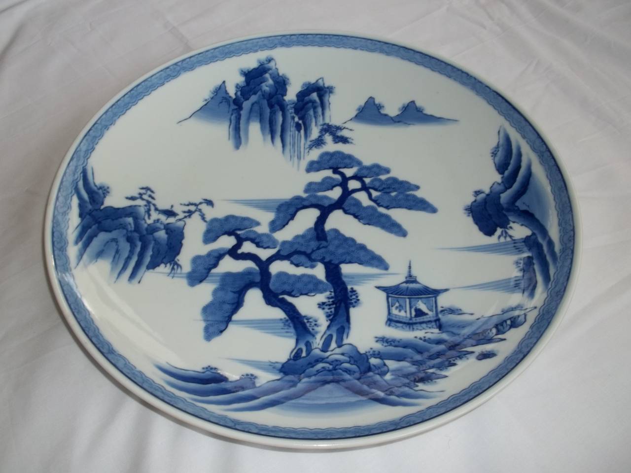 This is a very large and impressive Japanese porcelain blue and white charger of over 15 inch diameter.

The decoration shows a central large tree in a woodland setting with a pagoda all in different shades of under-glaze blue, with a border