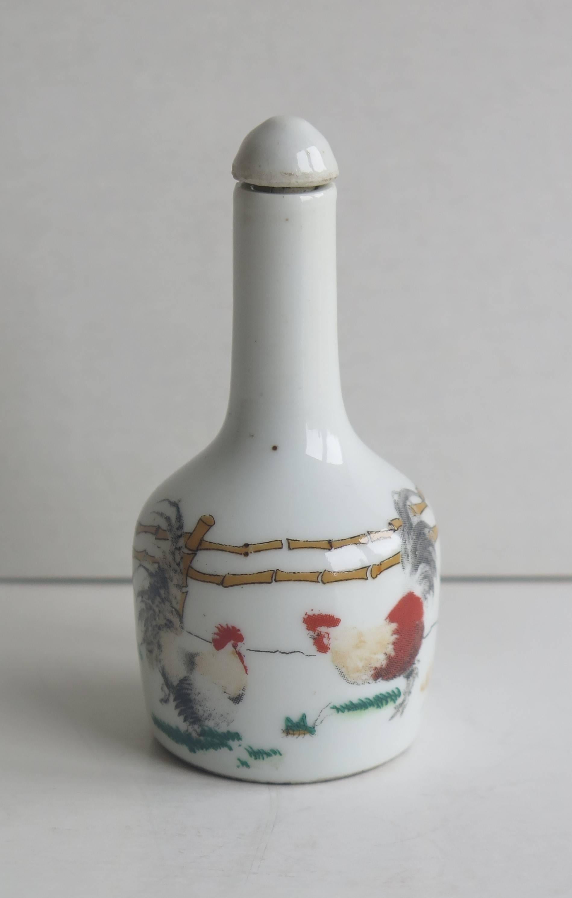 This is a good quality Chinese snuff bottle, made from porcelain, circa 1950. 

The cylindrical bottle has a tall narrow neck. The main decoration depicts two painted cockerels (or roosters) in a farm yard setting, with a fence and foliage. The