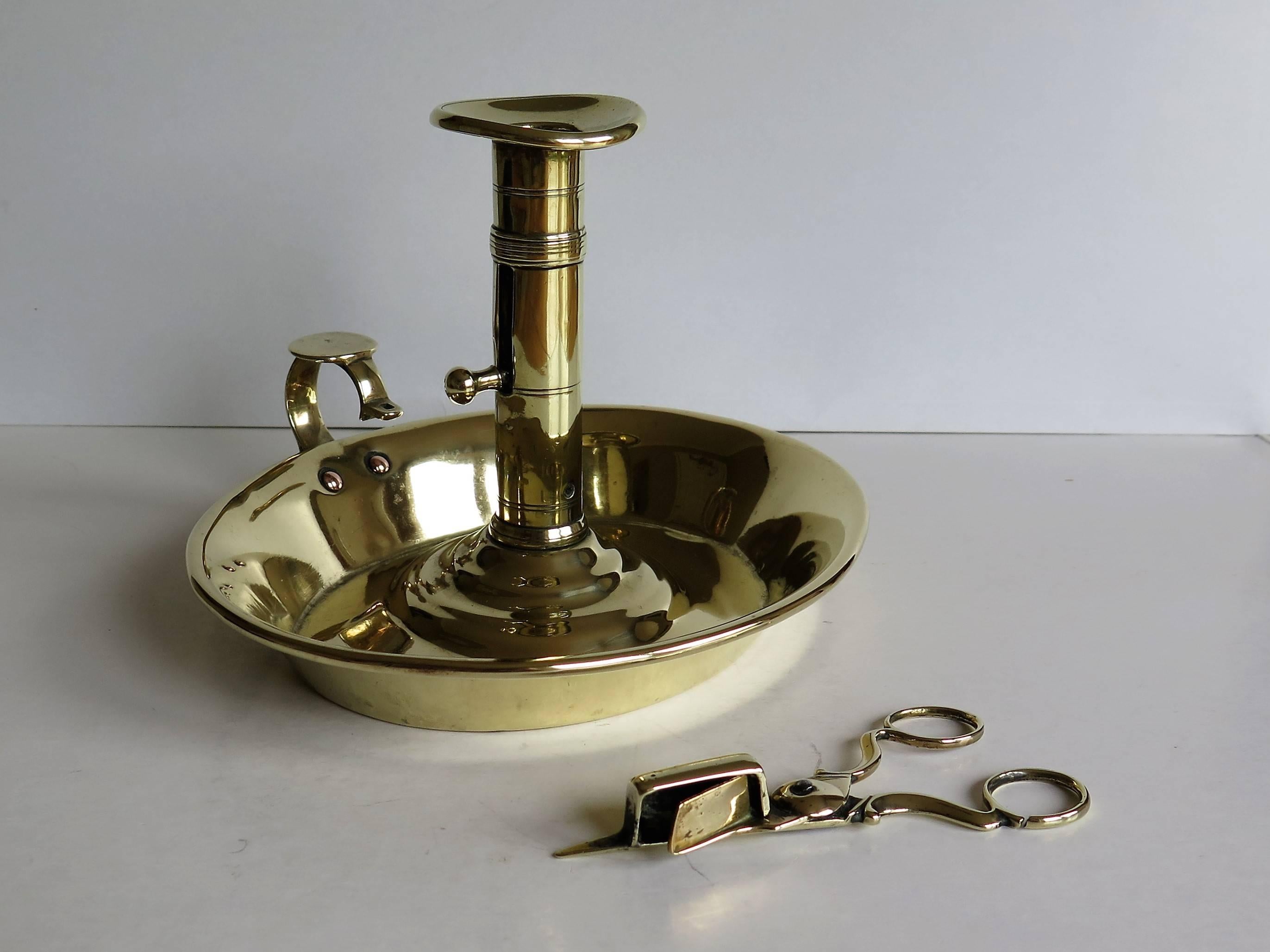 This is a late Georgian English chamberstick or candlestick with an associated wick snuffer or trimmer, both items made of brass and dating to the very early 19th century.

The chamberstick has an oval base with a high loop handle. The stem has a