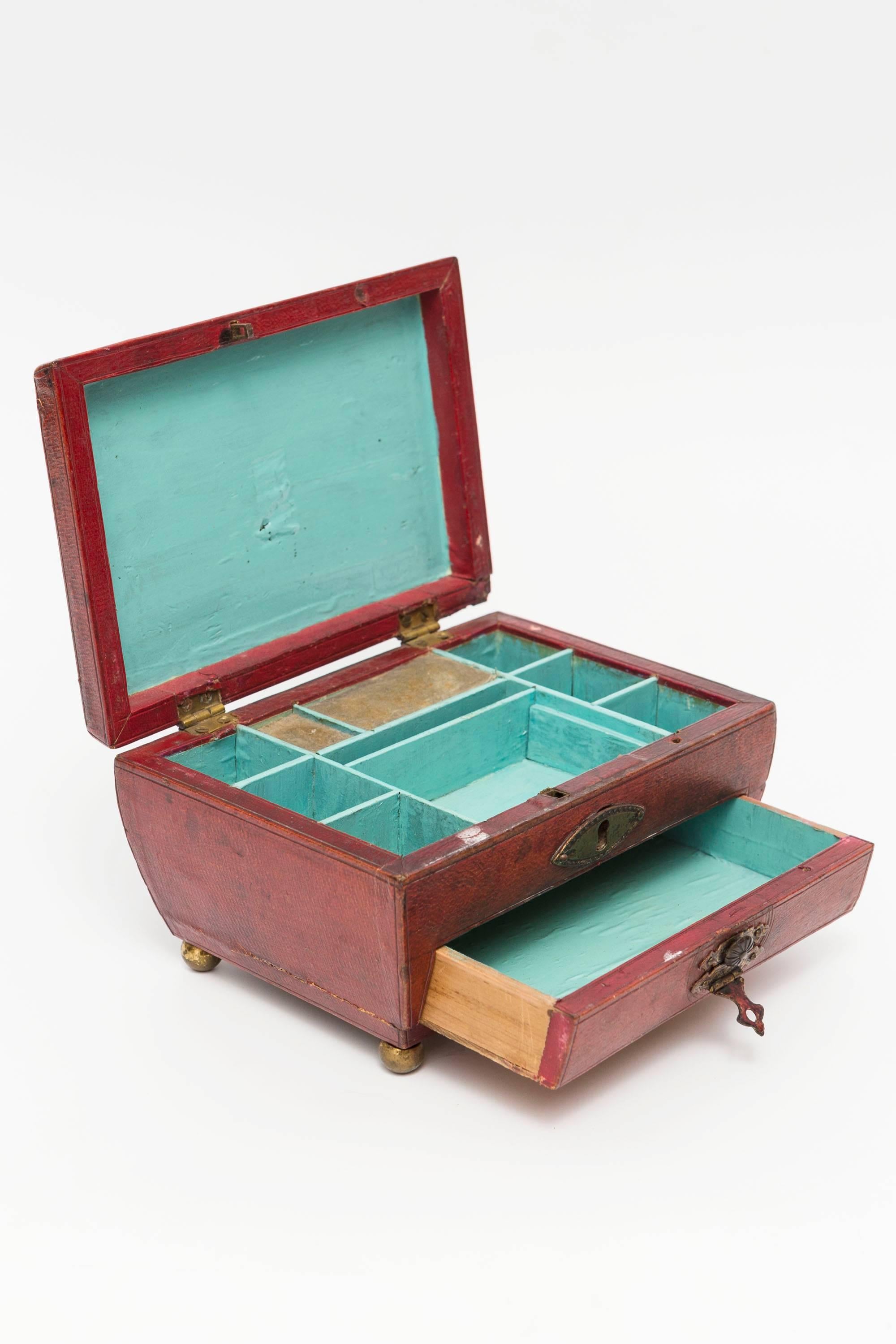 This is a 19th Century Moroccan red leather Georgian jewelry casket or box.  The top lid opens to reveal divided sections painted in a turquoise color. There is a small bottom drawer that pulls out with storage of single tray. All the hardware is