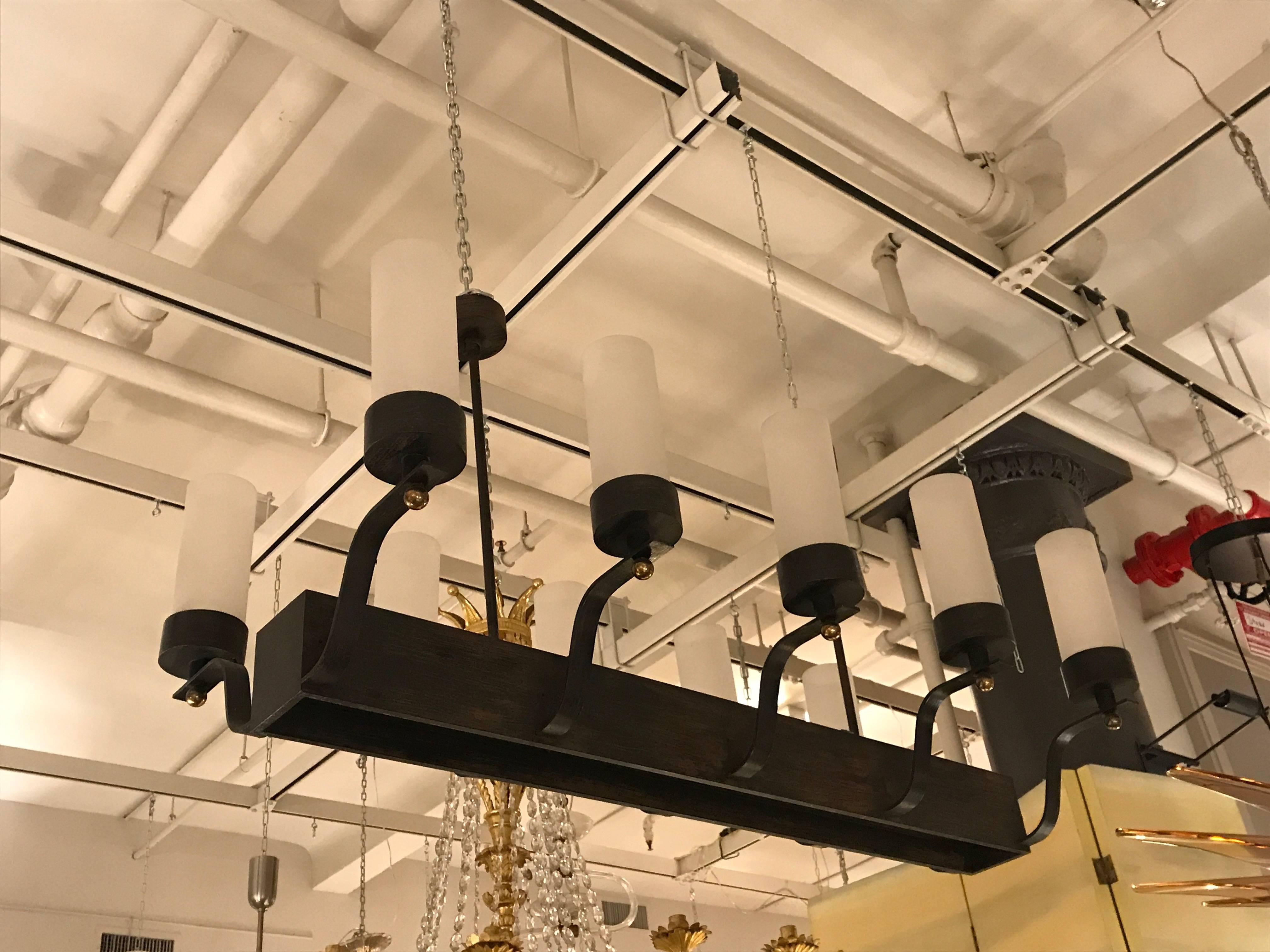 Patinated iron, rectangular structure with ten up-curving arms (five on each side) supporting cylindrical shades of white glass with gilded bronze, spherical finials. The iron body is inset with frosted glass panes for down light and hangs from two