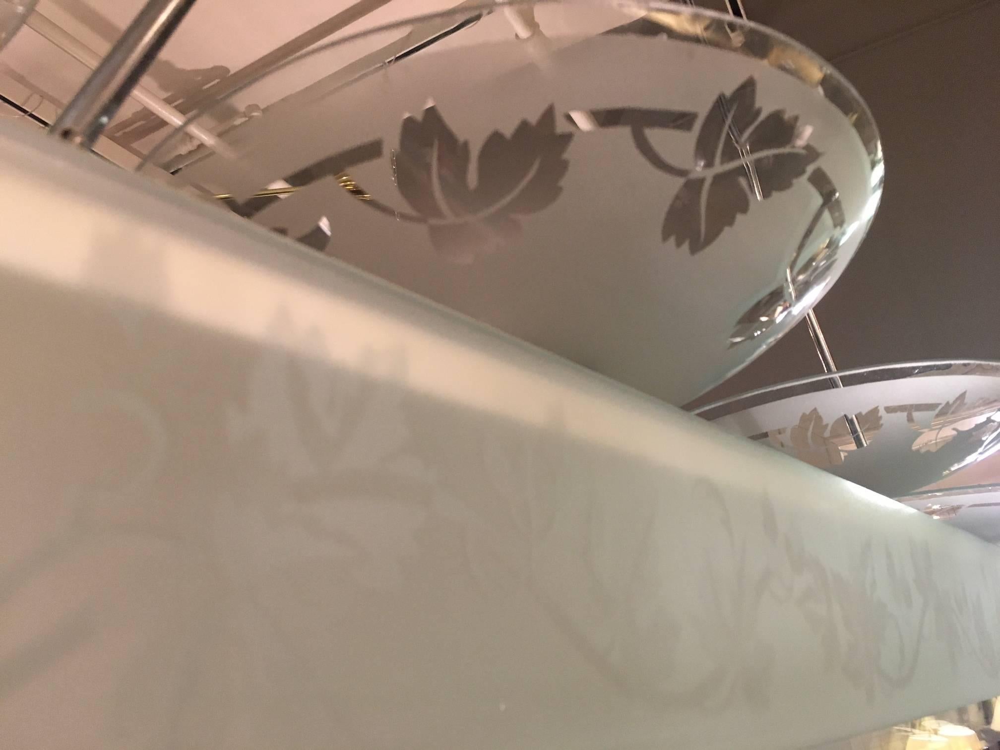 Vier Glasschalen, geätzt mit stilisierten Schwaden, montiert an einer langen, beleuchteten Chromstruktur mit ebenfalls geätzten Glaswänden, die vier Glaslamellen enthält und an zwei verchromten Stangen und einem Baldachin hängt.

UNSERE REFERENZ