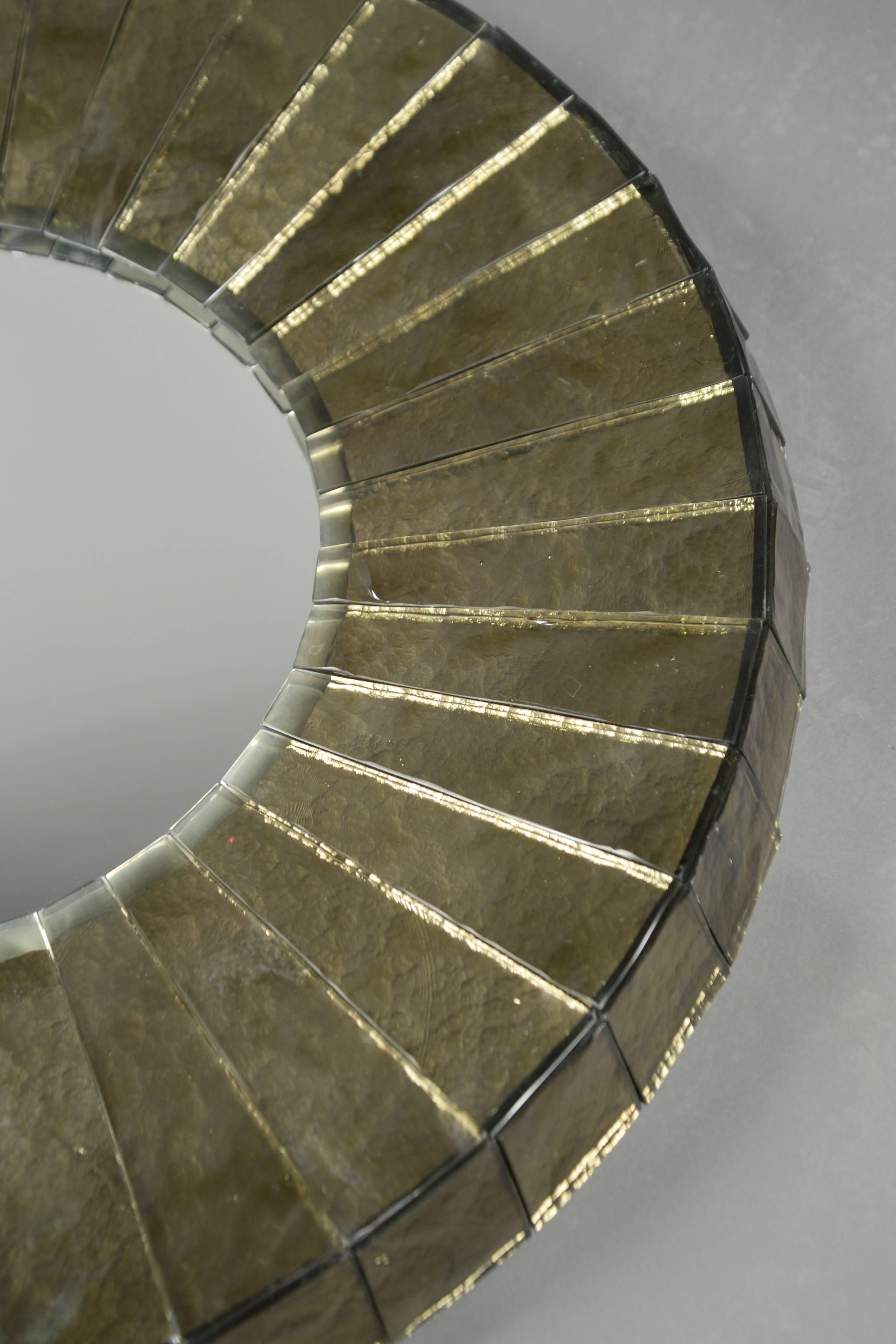 Hand-hewn wedges of thick mirrored gray glass, abutting similarly chiseled sides, encompassing the round mirrored glass plate. Model “Leucos”. The mirror plate itself is 17