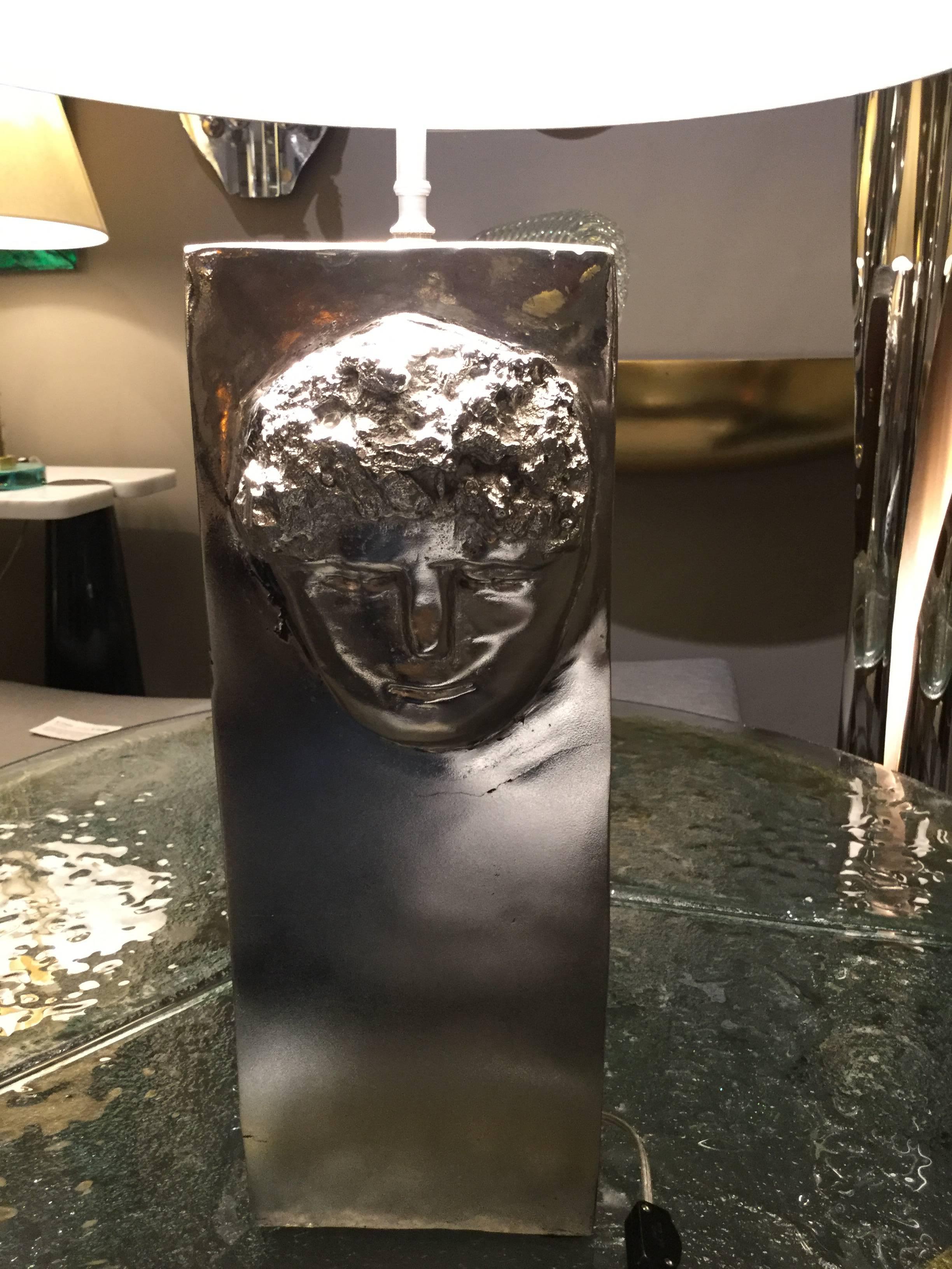 Cast aluminum cubic base, with a stylized visages facing forward and back. Inscribed signature on the base. 

(Lamp shade not included)