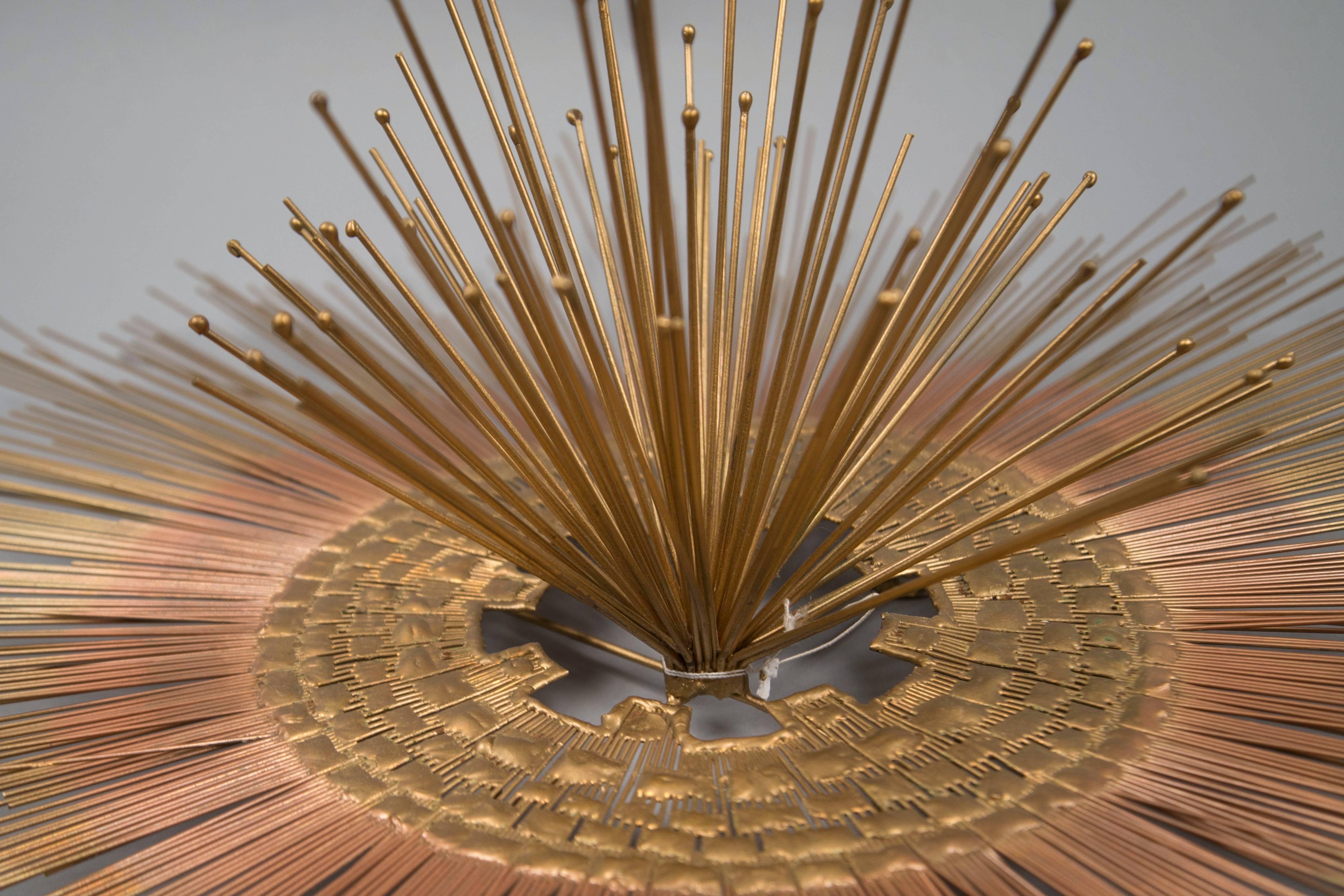 Brass wall sculpture featuring a copper-colored abstract ring around outward facing pins, sitting atop radiating brass rods of varying lengths.

Diameter: 30
