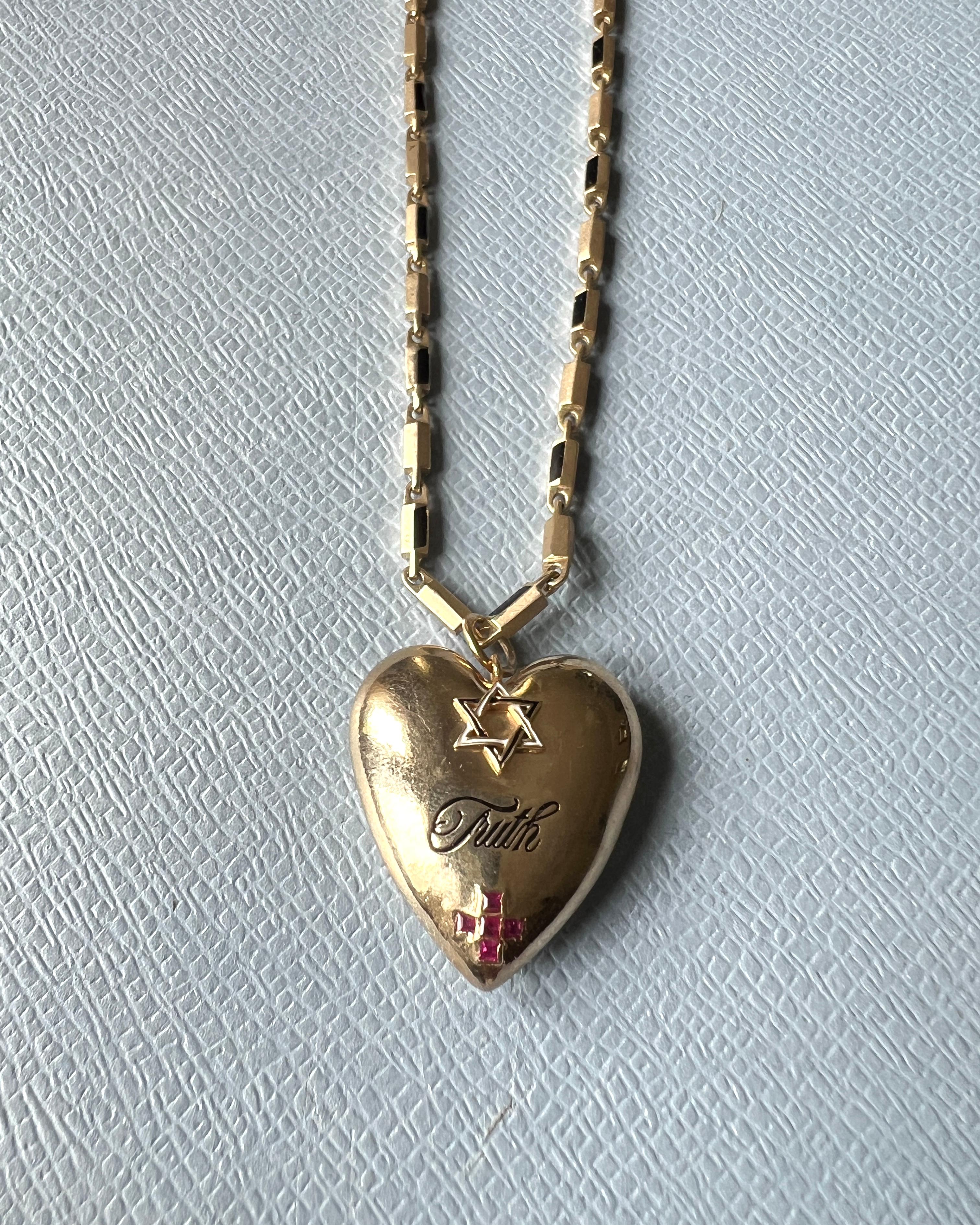 Este collar simbólico de J Dauphin tiene un colgante en forma de corazón de latón chapado en oro, con la palabra VERDAD grabada en el centro. Encima de la palabra hay una estrella de David, y debajo de ella, una cruz simétrica; cada símbolo tiene