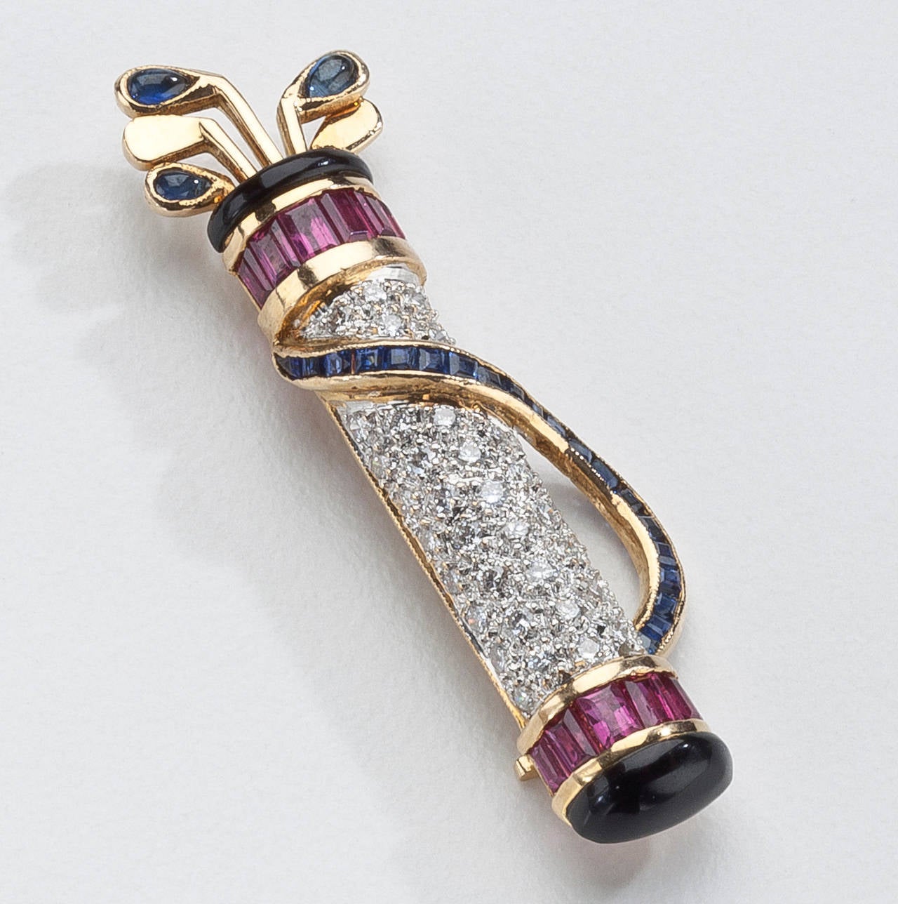 An 18 karat gold lapel brooch of a golf bag with five clubs.  The brooch is highlighted by twelve baguette cut rubies, sapphires, meli diamonds and onyx.  Signed Le Vian