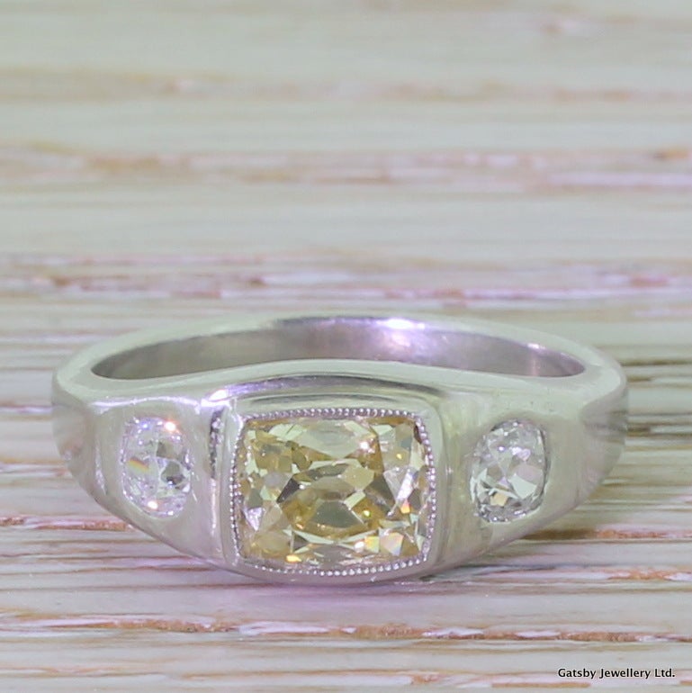 A glowing and unusual trilogy of diamonds in this gorgeous French Art Deco ring. The middle stone is a long cushion shaped old mine cut of a vibrant medium fancy yellow hue. Neatly set in a milgrained bezel setting and is mounted slightly higher