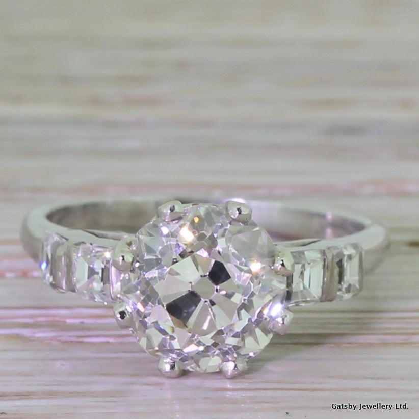 A mega and magnificent Art Deco treasure. This big, bright and vibrant old mine cut stone rests in a triumphant platinum setting. The central solitaire holds a soft champagne hue, deliberately emphasised by the four colourless asscher cut diamonds