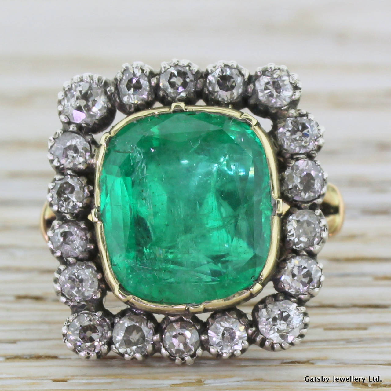 A Colombian emerald so bright and so green it looks electric. This grand cushion shaped stone is rub-over set in yellow gold with 18 white old mine cut diamonds set in silver in the surround. Set upon an elegant split shoulder band, this fine ring