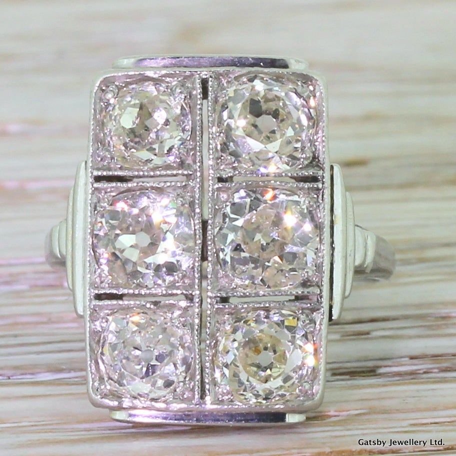 An immense and striking plaque ring set with six bright old mine cuts. Each of the vibrant diamonds is set within it’s own “box” in a pierced, milgrained and rigidly geometric setting. Step-down detailing on the shoulders leads to simple D-shaped