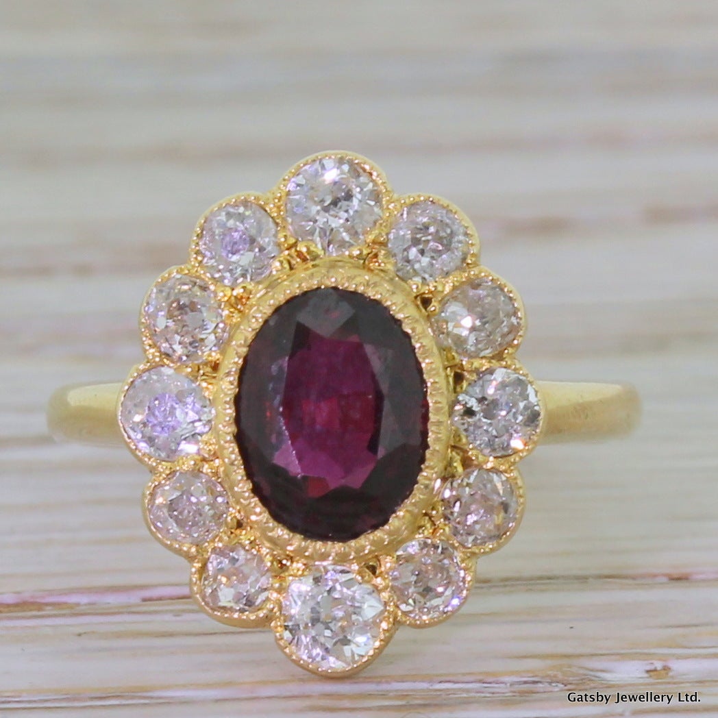 A fabulous ring; eye-catching and utterly arresting. An oval shaped ruby – natural and unenhanced – is rubover and milgrain set with twelve bright and lively old cut diamonds in the surround. The ruby is crimson with distinct flashes of fire,