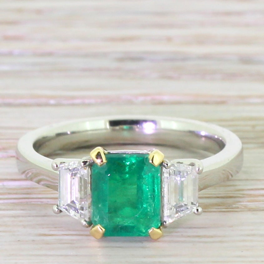Magnificence. An elegantly proportioned trilogy with a wonderful grassy green natural emerald cut emerald in the centre, flanked by a pair of clean ice-white trapeze cut diamonds. All in platinum with the exception of a the four 18k yellow gold