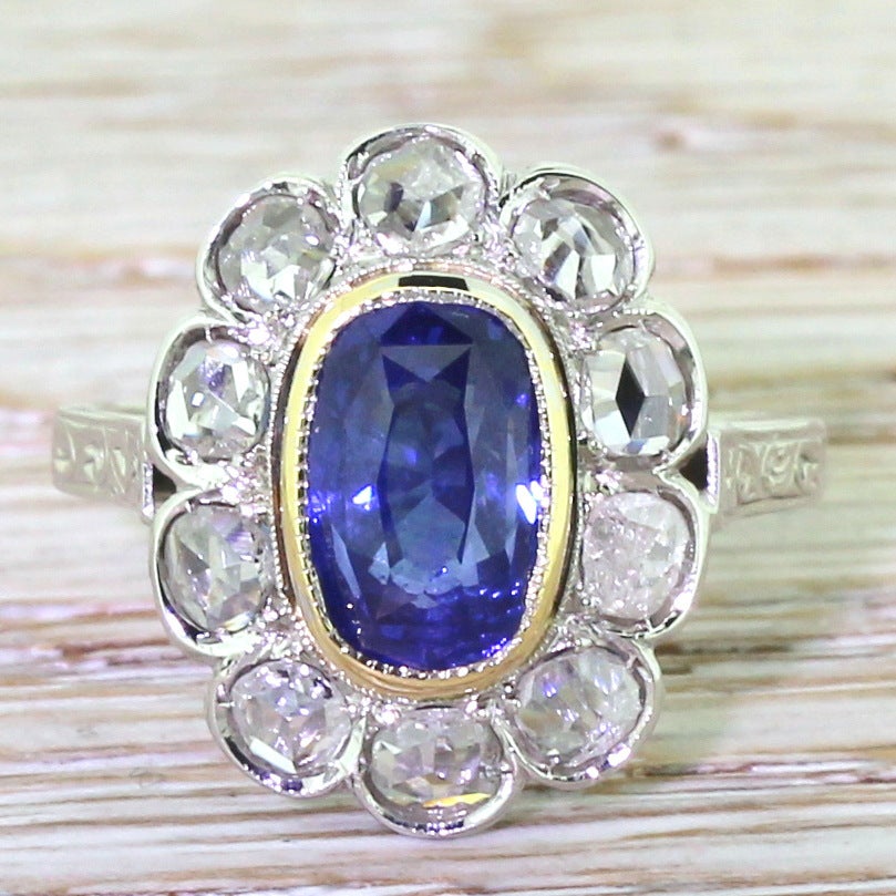 Impressive and indisputably stunning. Set with an old oval cut blue sapphire bezel set in 18k yellow gold,  surrounded by eleven high white and clean rose cut diamonds. A pierced and etched gallery leading to ornately engraved shoulders. A quite