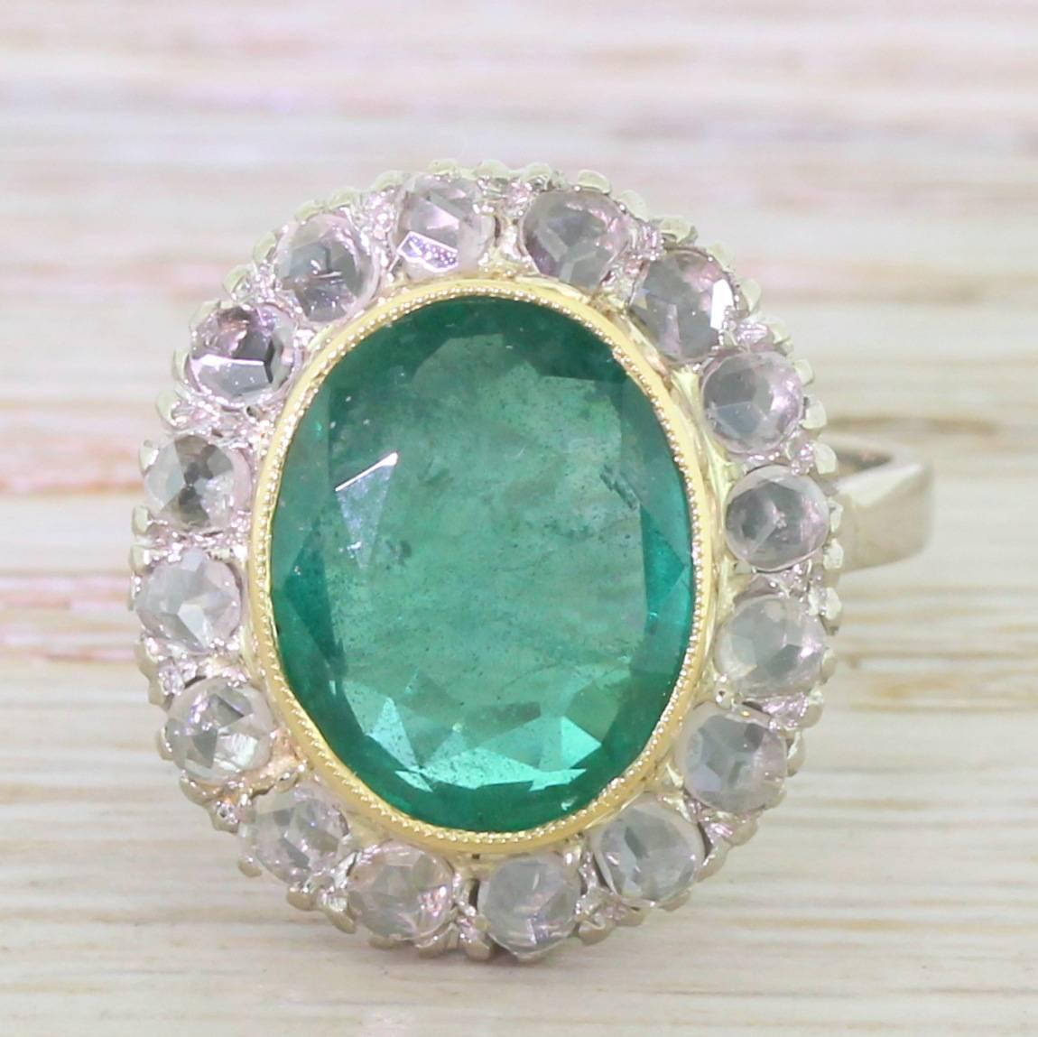 A bright, rich medium green oval cut emerald is rubover and milgrain set in yellow gold, with sixteen rose cut diamonds in the surround. This wonderful statement ring features fleur de lis shoulders at the shoulders leading to a flat white gold