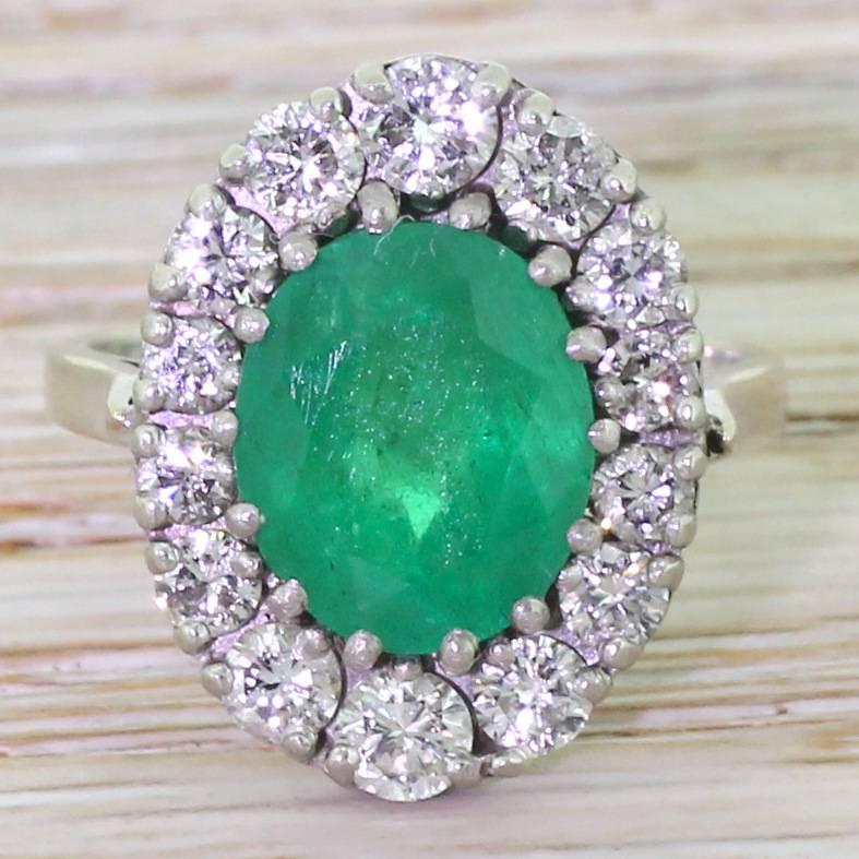 An emerald of the most intense and glowing green. This quite arresting stone is set in a surround of fourteen high white brilliant cut diamonds of graduating size atop a beautifully detailed hand crafted gallery. A ring that somehow becomes more