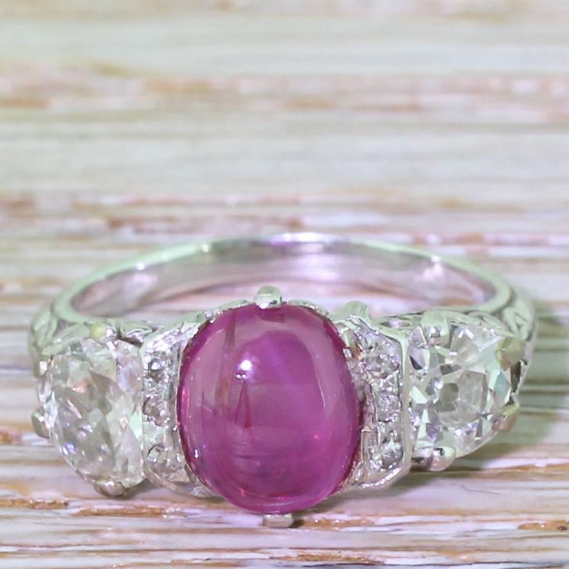 A captivating three stone ring. The glowing, pinkish red cabochon ruby (natural and unheated) is framed by two rows of eight-cut diamonds with a pair of high white old cut diamonds either side. The ornate detailing in the gallery continues into the