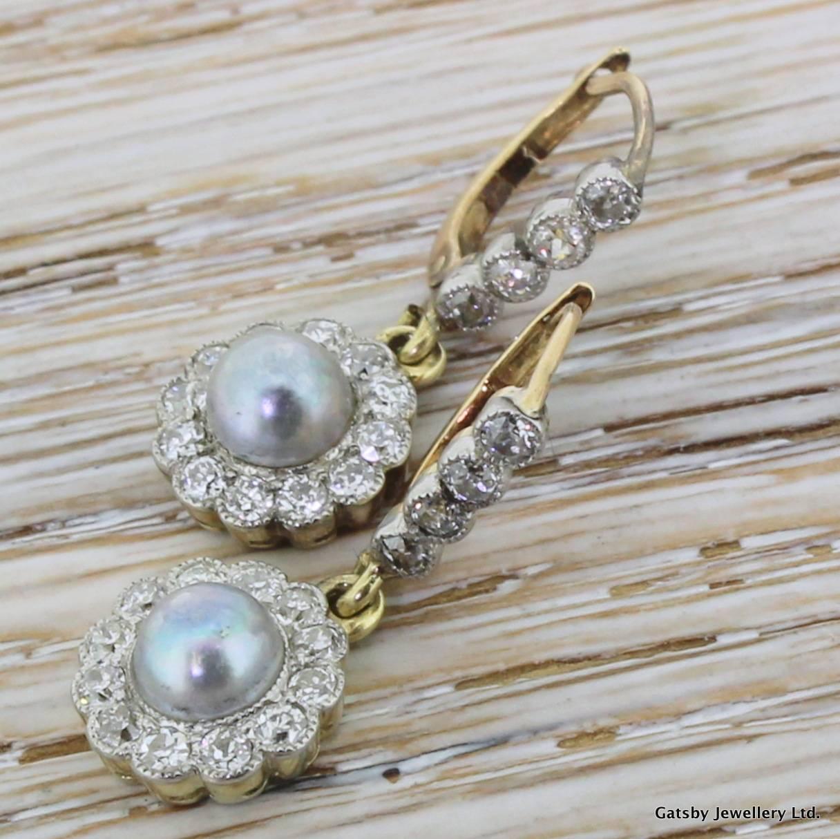 An adorable pair of earrings, set with lustrous grey pearls encircled by 12 high grade old cuts. The clusters dangle from a fixed bar which hold a further four diamonds. A gorgeous contrast between the grey pearl and the colourlessness of the