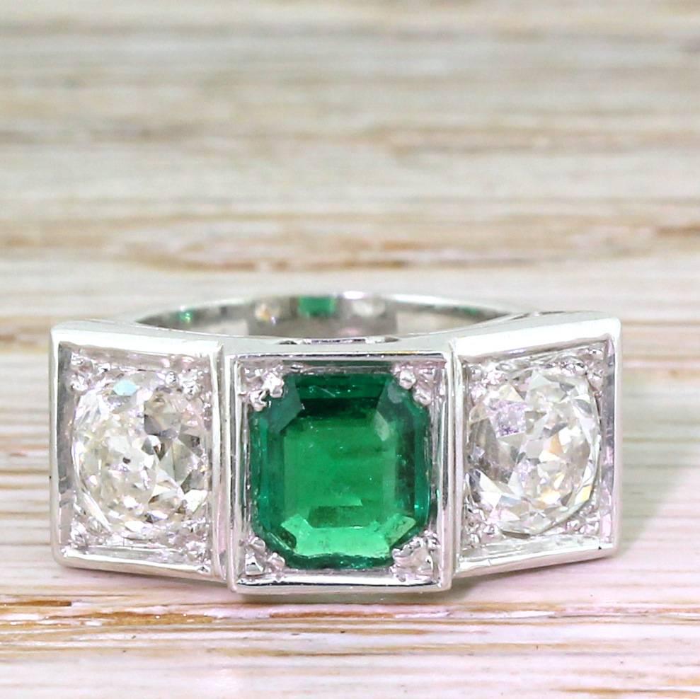 A proper ring, this. The emerald cut Colombian emerald in the centre is flanked by a pair of high white and very clean cushion shaped old cut diamonds. Each gem is set in its own box, within an unfussy open gallery leading to a substantial D-shaped