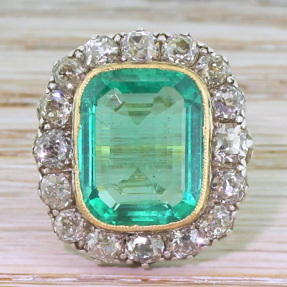 A hypnotically beautiful ring. The clean, clear and glowing Colombian emerald is rubover set in yellow gold, with sixteen cushion shaped old cut diamonds in the surround. The cluster sits nice and low to the finger, with incredibly detailed scroll