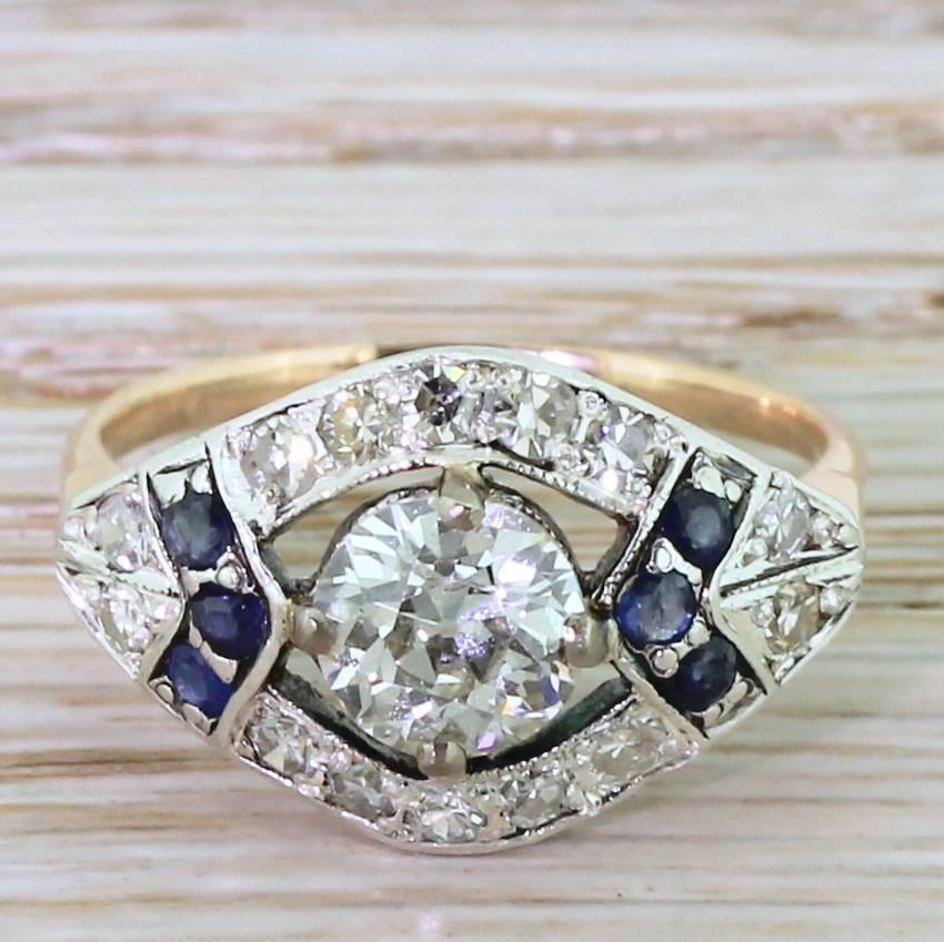 A flamboyantly beautiful ring. The centre stone – a 1.23 carat old European cut displaying impressive fire and brilliance – “floats” within a lateral marquise set with fourteen eight-cut diamonds and six natural, blue sapphires. All the stones are
