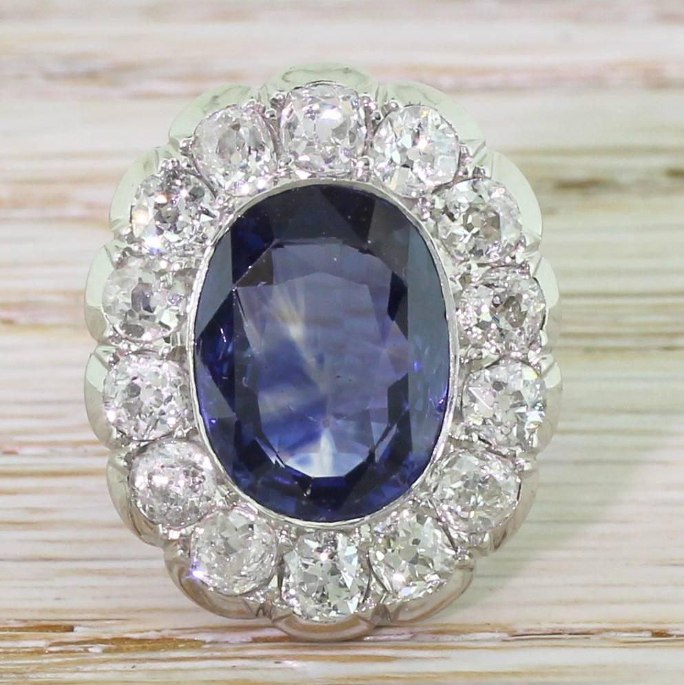 A grand and impressive statement of a ring. The natural and unheated Ceylon sapphire is deep, glowing blue with clear flashes of indigo. The centre stone is surrounded by fourteen large and white old cut diamonds in cutaway settings. Beautiful