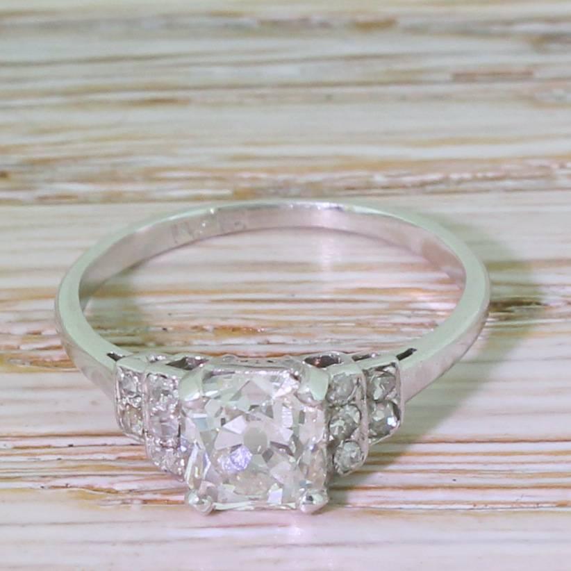 Pure Deco class. The square cushion shaped old cut diamond in the centre is glowingly white, internally clean and bursting with life and fire. This quite exceptional stone is secured in a four claw collet, with step-down shoulders featuring ten