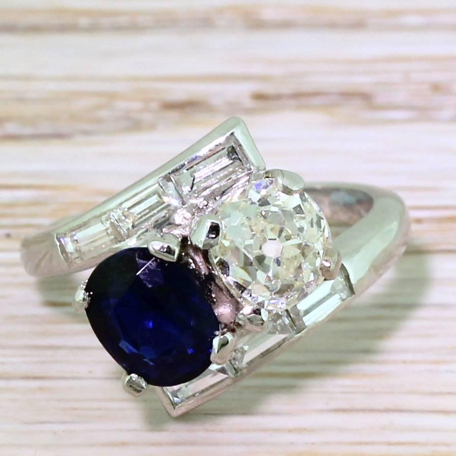 Wonderful. This daring crossover ring features a cushion shaped old cut diamond,  the soft champagne hue  ofwhich contrasts with a deep, rich tone of the natural blue sapphire. Each stone is secured in a four claw collets nestled within a platinum