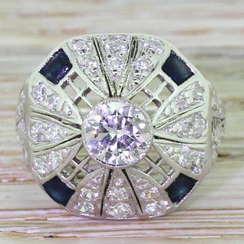 A late Art Deco ring in platinum that is absolutely bursting with originality and intrigue. The central old European cut diamond – high white and full of vibrancy – is rubover set and floating within a mount that holds an incredible amount of