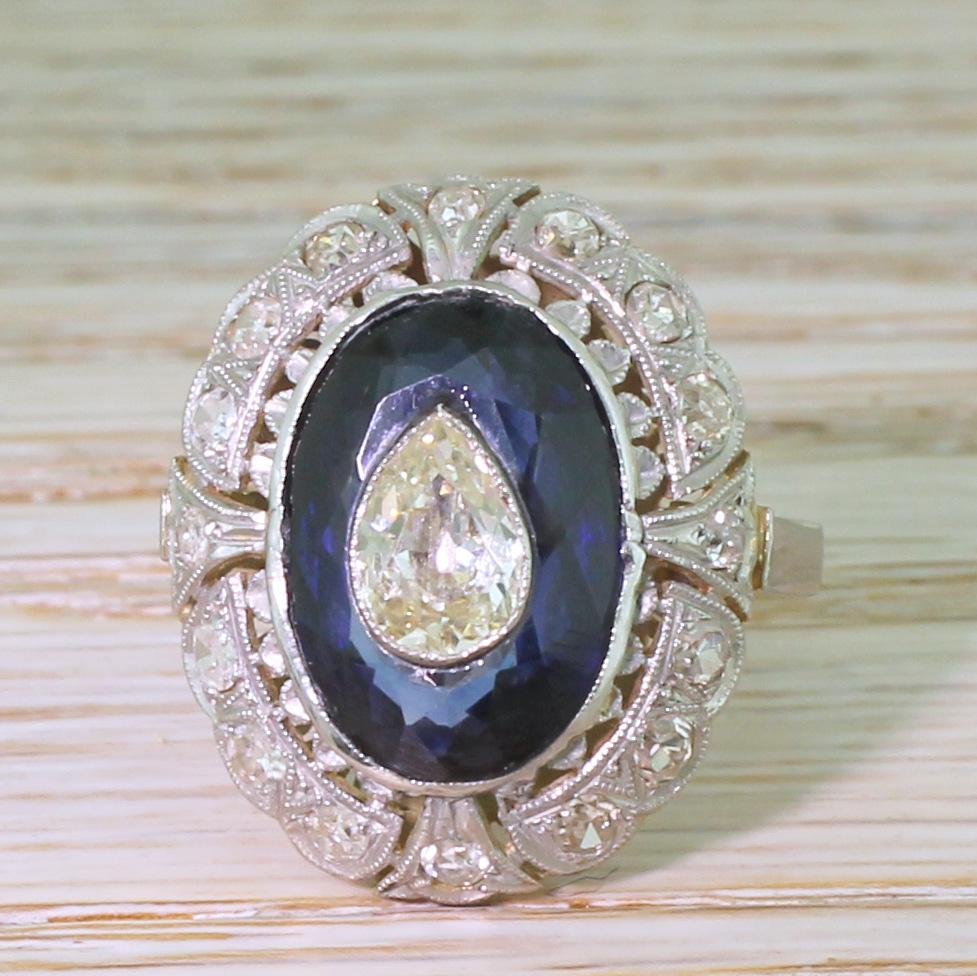 What a wild and wonderful piece! The fantastic ring features an old pear cut diamond, rubover and milgrained, and set in the middle and within a natural, deep blue sapphire. The finely pierced and crafted surround features sixteen white and bright