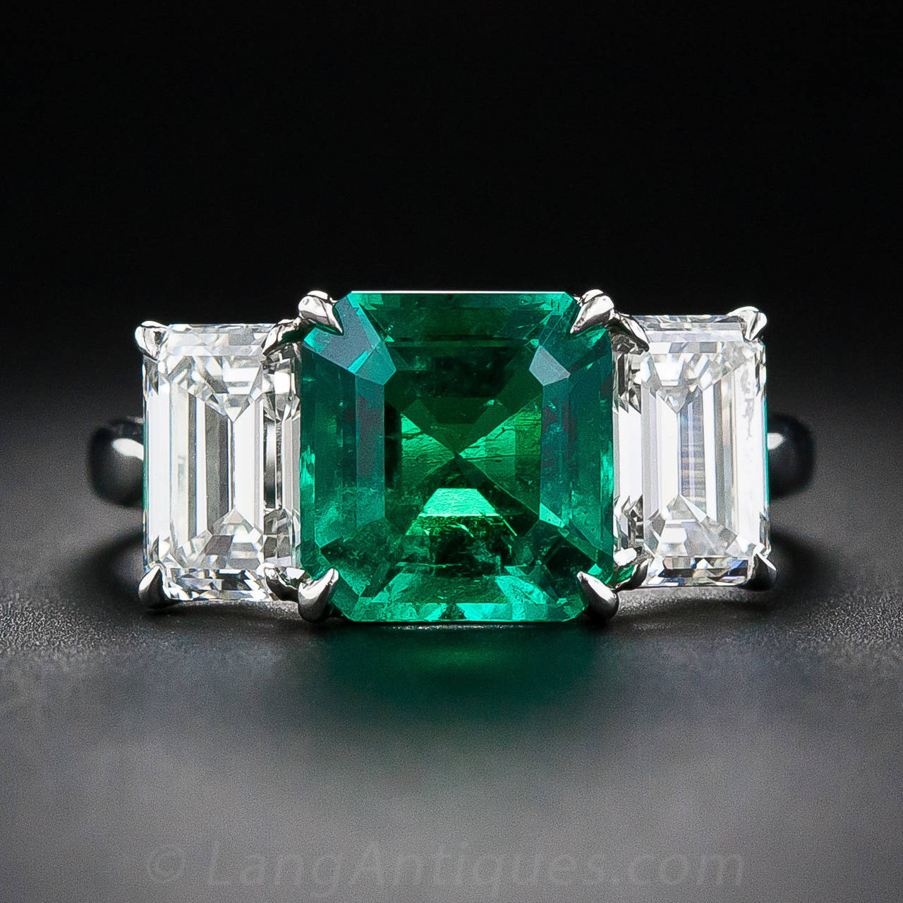 A gorgeous, richly saturated, vibrant green and crystalline square emerald-cut emerald - weighing 2.75 carats - radiates from between a pair of bright-white, (1.11 and .96 carats respectively), emerald-cut diamonds in this classic and captivating