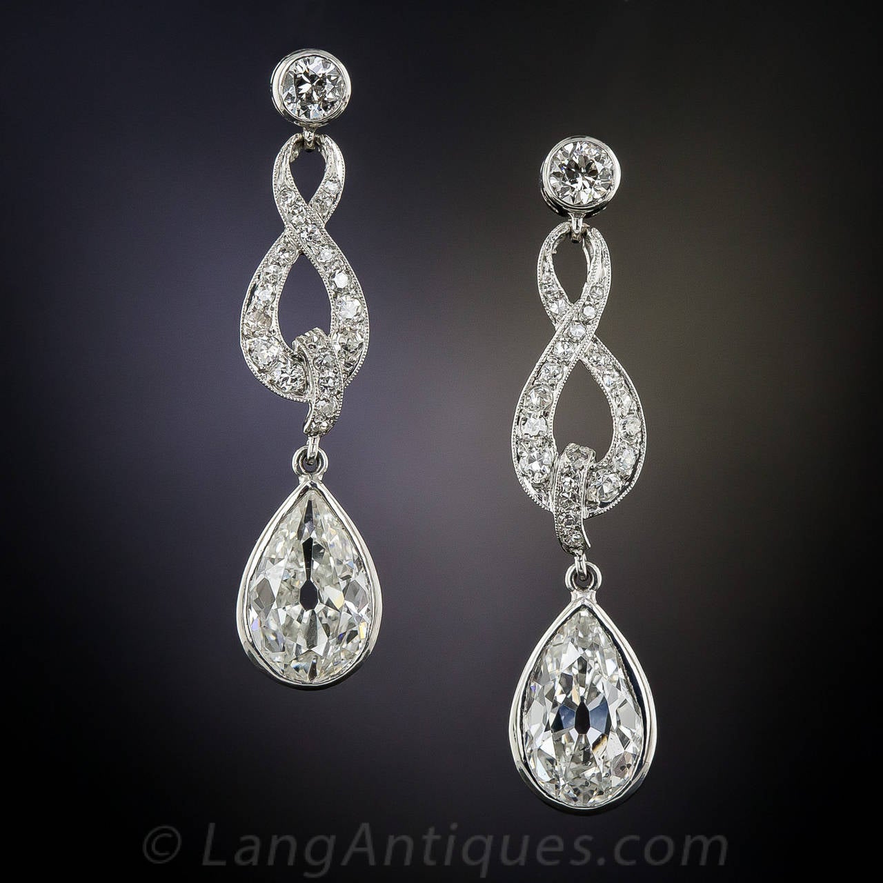 A sparkling pair of old mine-cut pear-shape diamonds, totaling approximately 2.65 carats, swing and sway from within bezel settings surmounted by beautifully stylized figure-eight, or infinity motifs, in these delightful and dazzling drop earrings