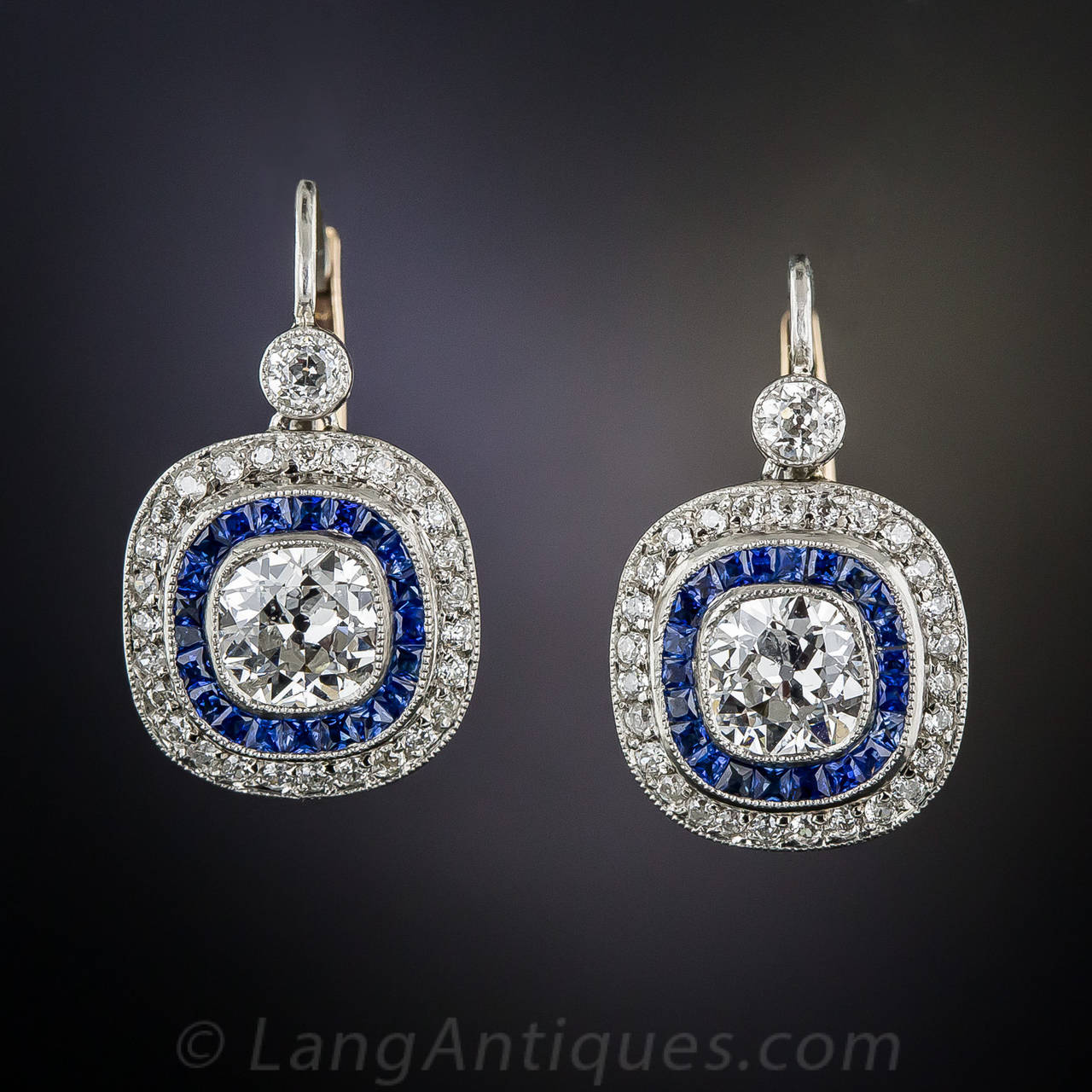 A beautiful pair of bright white antique cushion-cut diamonds, weighing one carat-plus each, have found a perfect new home in these exceptionally well made earrings, rendered in platinum, diamonds and electric blue, faceted calibre sapphires. The