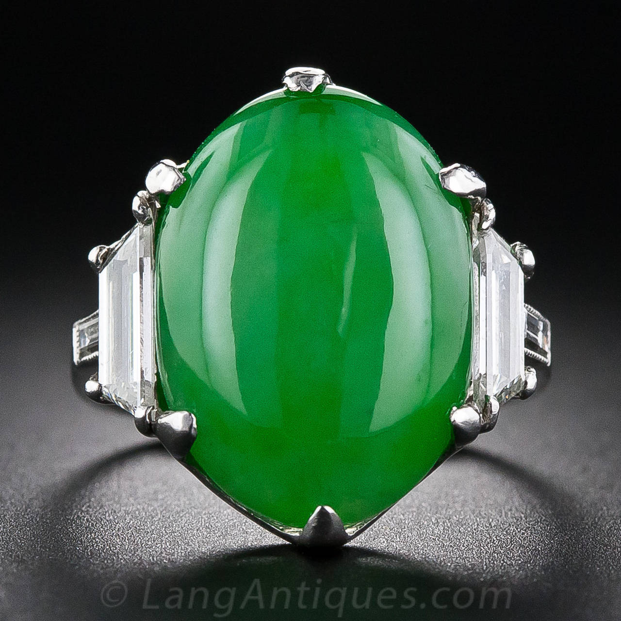 An impressive Art Deco ring set with a jadeite cabochon of vibrant green, accompanied by a GIA certificate stating natural color, flanked by two elegant trapezoid diamonds with baguette diamonds on the shoulders. The handmade platinum setting is